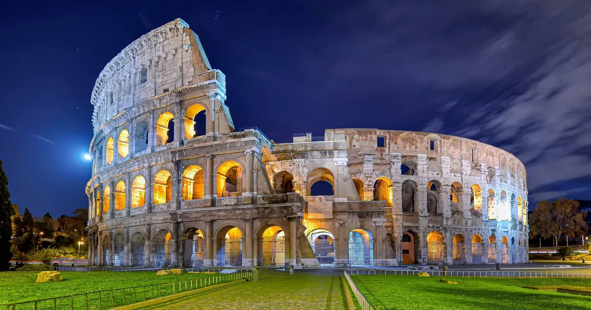 The Colosseum floodlit at night under a deep blue sky, its arches glowing gold above a green lawn