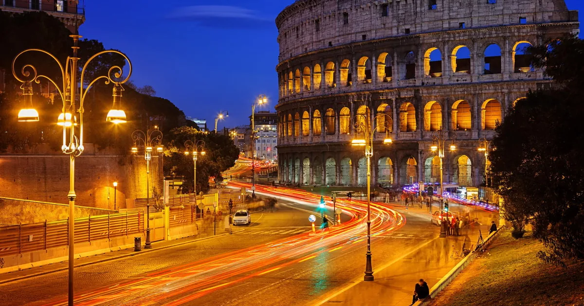 The Colosseum at dusk above a curving street with light trails from traffic and ornate street lamps