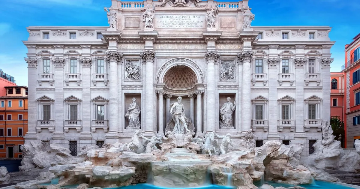 The full Trevi Fountain facade with its statues, carved rocks and turquoise basin under a bright blue sky, framed by red and ochre buildings