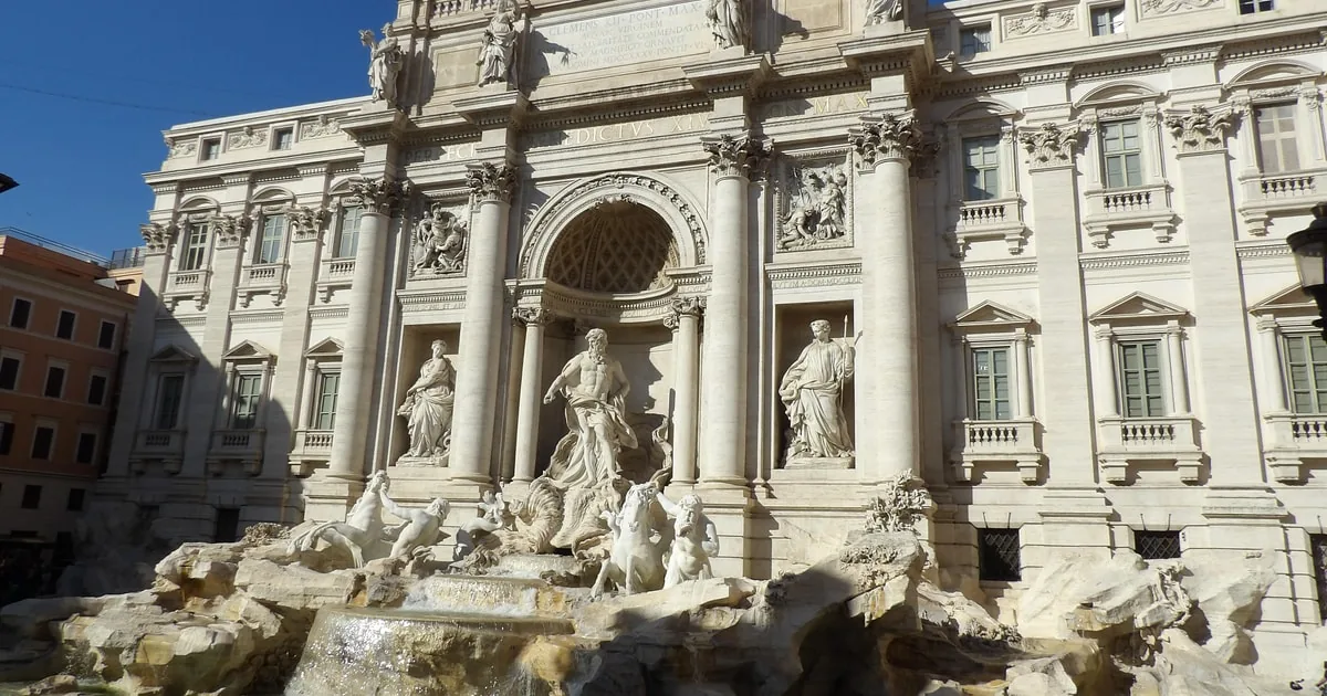 The Trevi Fountain's facade and statues in bright sun under a blue sky, the lower cascade in shadow