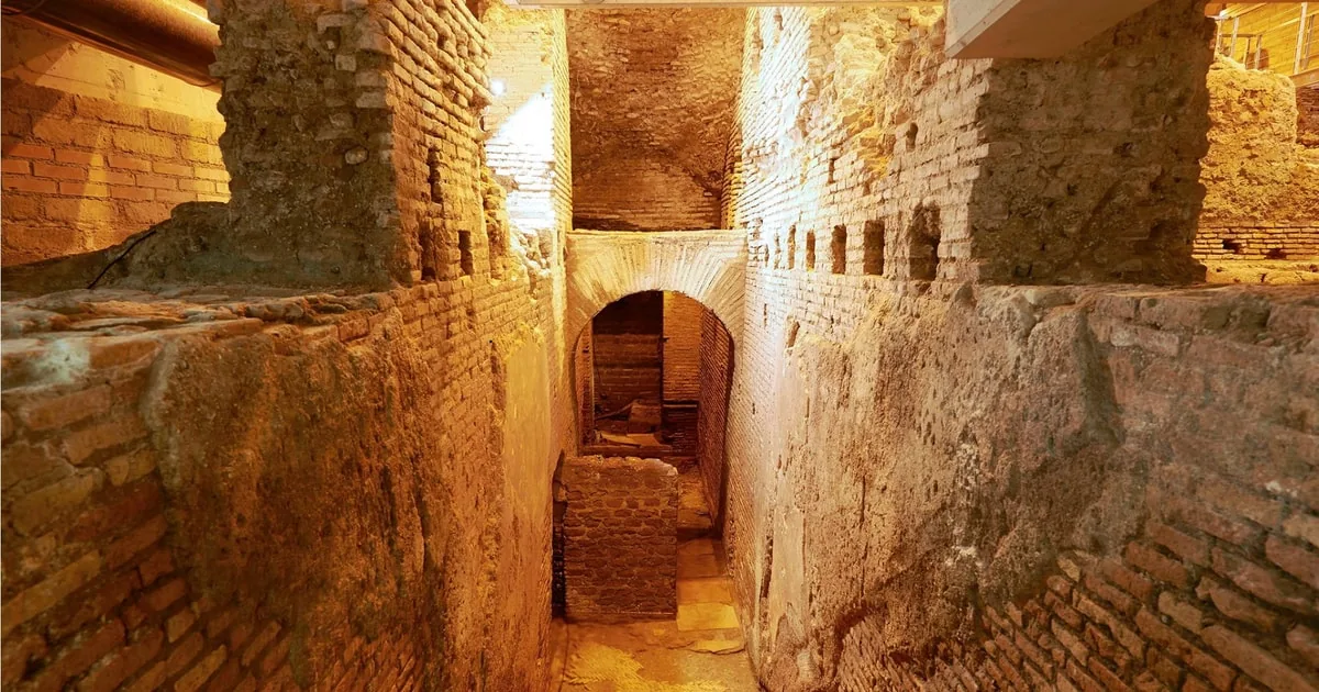 A narrow passage between tall old brick walls leading to a low arch, lit in warm orange light
