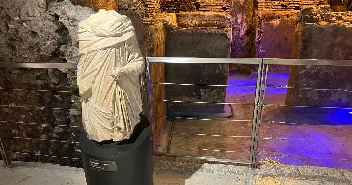 A headless marble statue in draped robes on a black plinth beside a metal railing in a warmly lit underground excavation